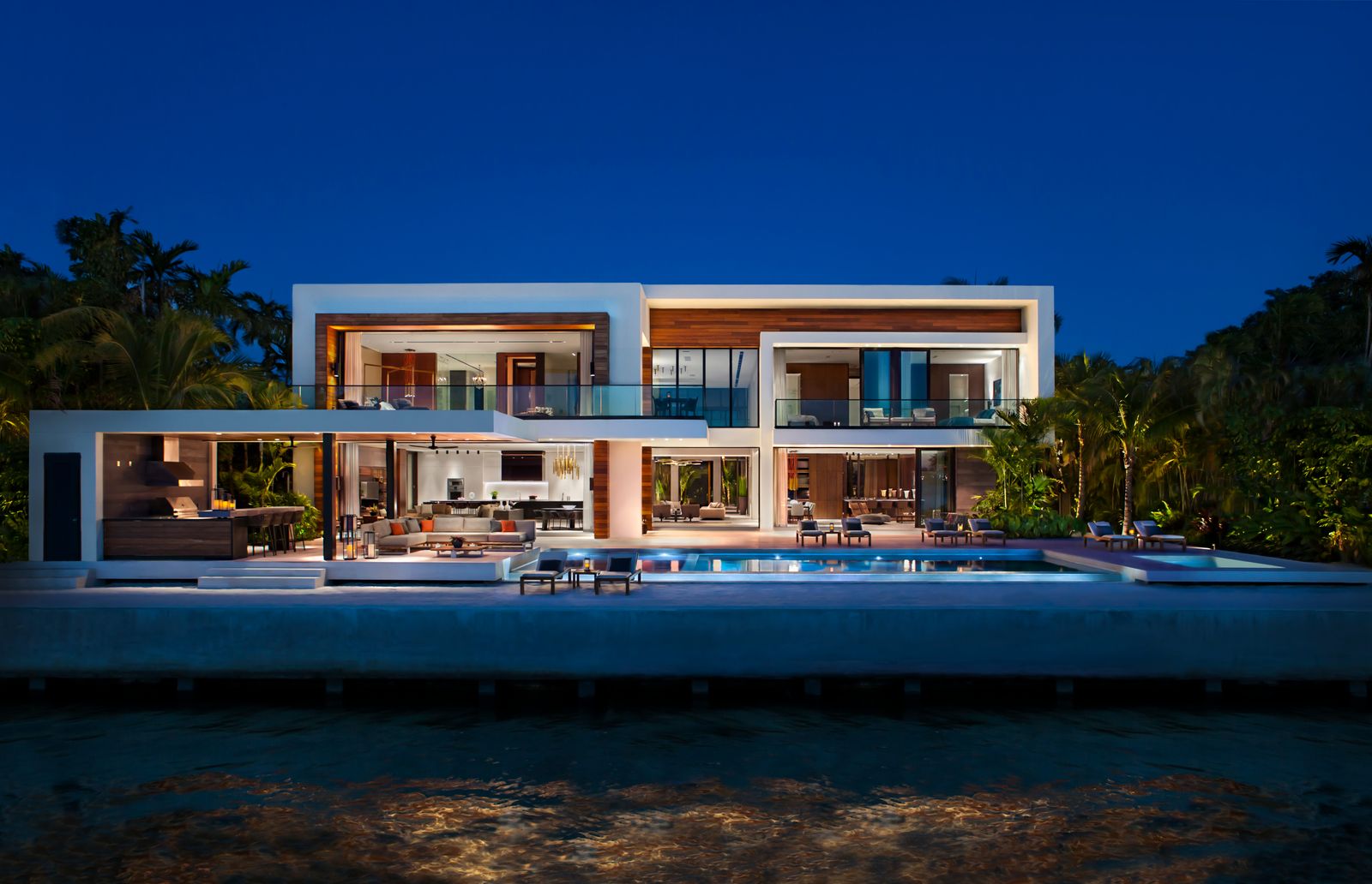 Modern waterfront home by Bart Reines Luxury Homebuilder at dusk, with a lit pool and open lower level facing the water