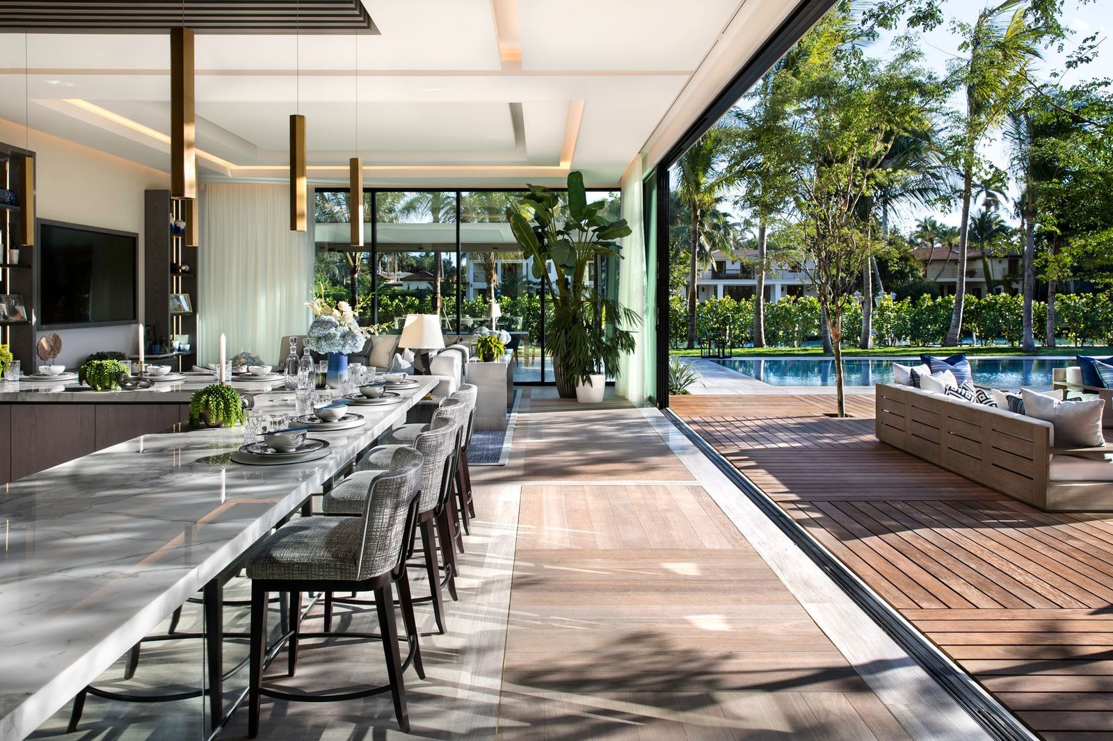 Indoor-outdoor kitchen with a long marble island and bar seating that opens onto a wood deck, pool and garden