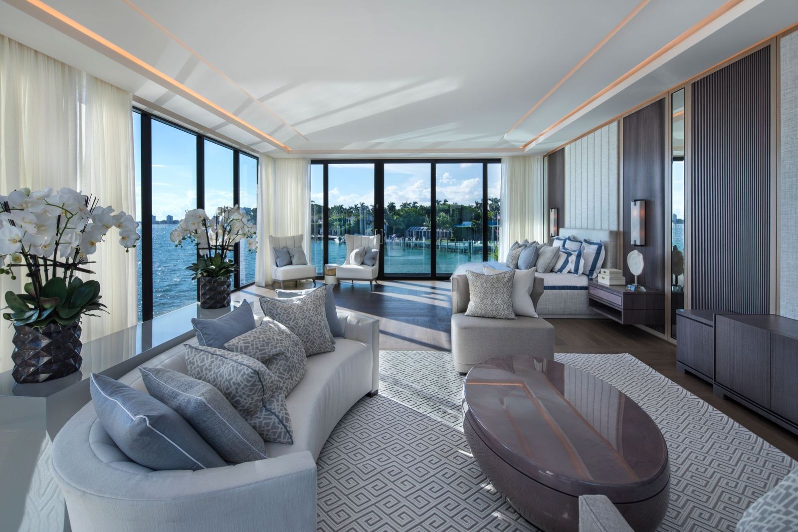 Primary suite sitting area with a curved sofa, orchids and floor-to-ceiling windows framing water views