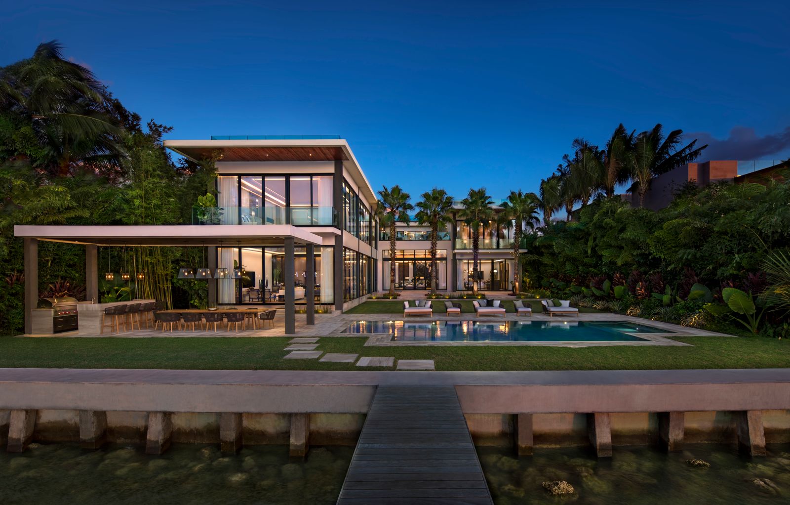 Rear of a Bart Reines waterfront estate at dusk: lawn, pool, outdoor kitchen pavilion and a dock walkway to the seawall