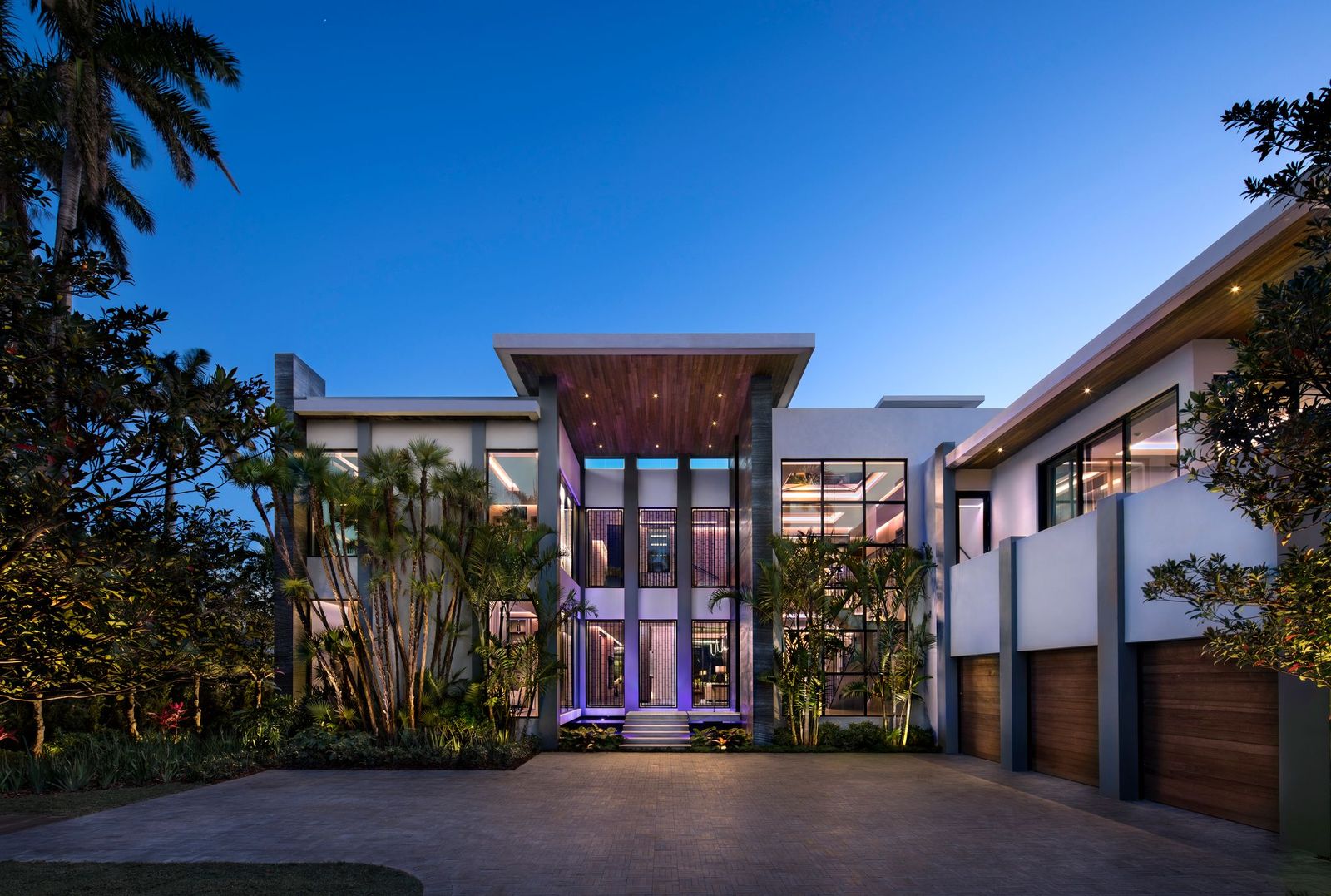Front facade of a Bart Reines custom home at twilight, with a double-height glass entry, wood soffit and three-car garage