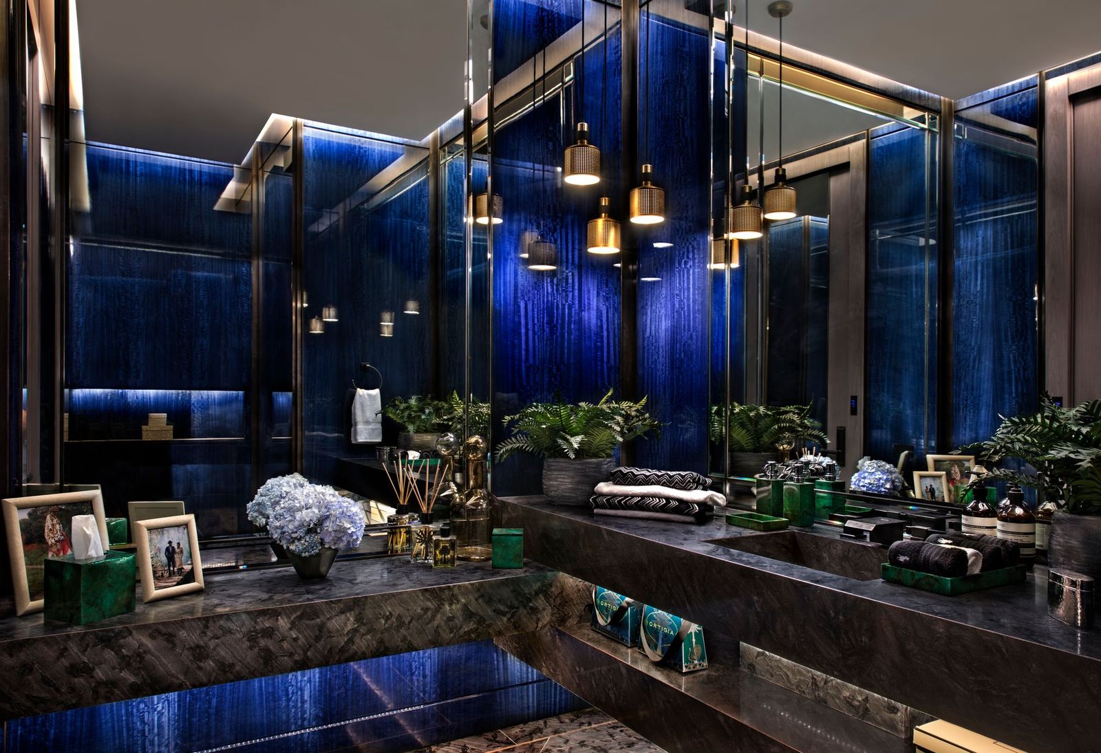 Powder room with backlit blue glass walls, a dark stone vanity and brass pendant lights